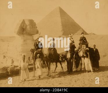 Trois femmes et un homme en tenue occidentale, assis sur des chameaux, plusieurs hommes locaux se tiennent devant les chameaux tenant les rênes, une pyramide et le sphinx en arrière-plan. Banque D'Images