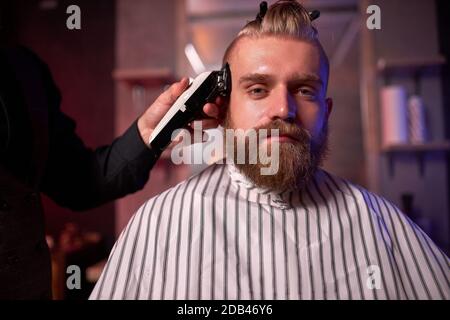 confiant caucasien barber mster coupe les cheveux et la barbe des hommes dans le salon de coiffure, professionnel coiffeur fait la coiffure pour un jeune homme, il travaille avec la tondeuse Banque D'Images