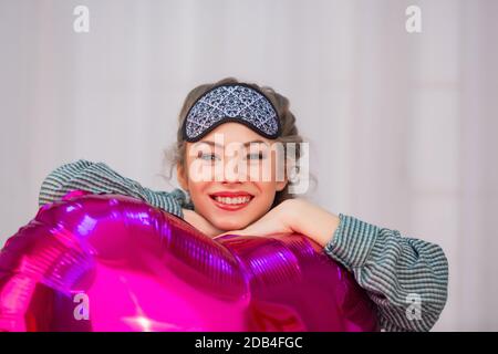 Jeune femme dans le masque de sommeil câlins rose coeur en forme de ballon et sourit Banque D'Images