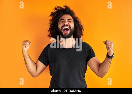 joyeux jeune indien ou arabe célébrant surpris et surpris par le gagnant de gagner, le concept. gai gars avec de longs cheveux sombres brillent avec bonheur Banque D'Images