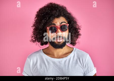 portrait de l'homme à barbe mauriquement dans des lunettes de soleil isolé sur fond rose, jeune homme en t-shirt blanc a l'air tendance, élégant Banque D'Images