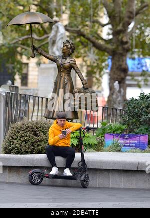 Londres, Angleterre, Royaume-Uni. Licester Square: Jeune homme avec un e-scooter sur son téléphone portable devant une statue de Mary Poppins Banque D'Images