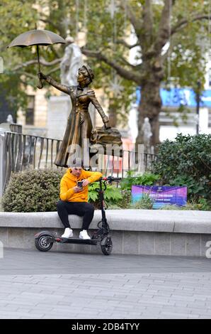 Londres, Angleterre, Royaume-Uni. Licester Square: Jeune homme avec un e-scooter sur son téléphone portable devant une statue de Mary Poppins Banque D'Images