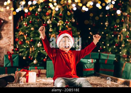 Le garçon a un chapeau de père noël sur sa tête. En arrière-plan, il y a un grand arbre de Noël décoré avec des cadeaux en emballage vert et un noeud. Noël Banque D'Images