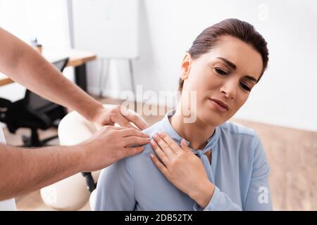 Masseuse massant l'épaule de la femme d'affaires assise sur une chaise de massage au bureau sur un arrière-plan flou Banque D'Images