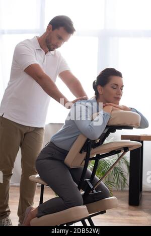 Masseuse avec des mains étirées massant le dos de la femme souriante avec les yeux fermés sont assis sur la chaise de massage dans le bureau Banque D'Images