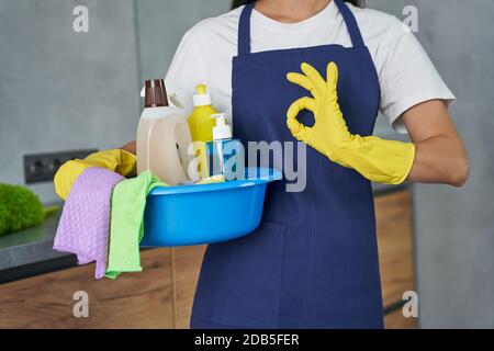 Photo courte de femme de ménage montrant le panneau ok tout en tenant le contenant plein de produits et d'équipements de nettoyage, debout dans la cuisine moderne. Ménage et entretien ménager, concept de service de nettoyage Banque D'Images