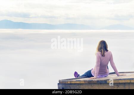 Femme en vêtements de sport reposant sur une plate-forme en bois et profitant de la vue de la vallée couverte de brouillard en dessous. Randonnée, réalisation, attente, optimisme an Banque D'Images