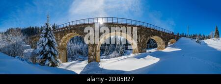 Viaduc de pierre (pont d'arche) sur le chemin de fer à travers la forêt de sapins enneigés de montagne. La neige dévie sur le côté de la voie et le givre sur les arbres et les fils électriques. Banque D'Images