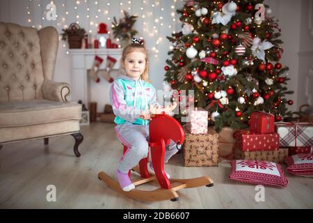 Une petite fille dans un survêtement est assise sur un fauteuil à bascule en bois rouge orignal près d'un arbre de Noël. Décorations de Noël, nouvel an, fêtes Banque D'Images