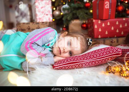 Une petite fille dans les vêtements de la maison est couché sous l'arbre de Noël près des boîtes avec des cadeaux sur l'oreiller et des rêves. Lumières des guirlandes, le befo de nuit Banque D'Images