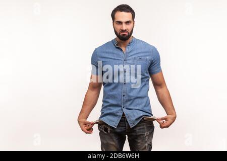 Pauvre homme contrarié avec la barbe dans des jeans et une chemise bleue montrant des poches vides regardant l'appareil photo avec tristesse, faillite, problèmes financiers. Intérieur s Banque D'Images