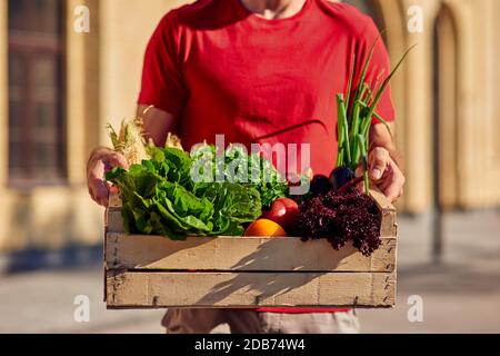 Plan court d'un messager masculin en uniforme tenant une boîte en bois avec une épicerie verte fraîche tout en se tenant sur une rue ensoleillée. Concept de livraison de nourriture Banque D'Images