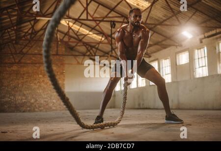 Homme fort et masculin utilisant des cordes d'entraînement pour l'exercice à la salle de gym à l'intérieur de l'ancien entrepôt. Un athlète qui s'efforce avec un cordage de combat. Banque D'Images