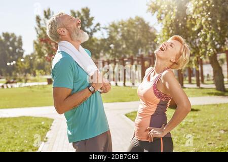 Être en forme. Joyeux couple sportif senior s'amusant en se reposant après l'entraînement, en parlant, debout dans le parc à l'extérieur. Des personnes actives faisant du sport Banque D'Images