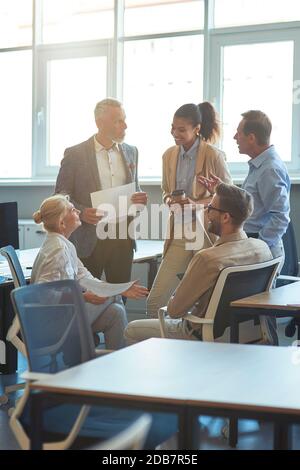 Photo verticale de gens d'affaires multiraciaux gais ayant une réunion dans le bureau moderne, discutant du travail et souriant Banque D'Images