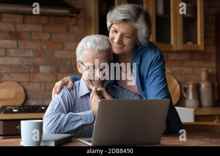 Couple mûr souriant qui se serre, utilisant un ordinateur portable dans la cuisine ensemble Banque D'Images