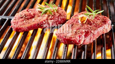 Deux steaks de bœuf cru épais, tendres et juteux, garnis de grillades de romarin sur les coals chauds glacés d'un barbecue en gros plan Banque D'Images