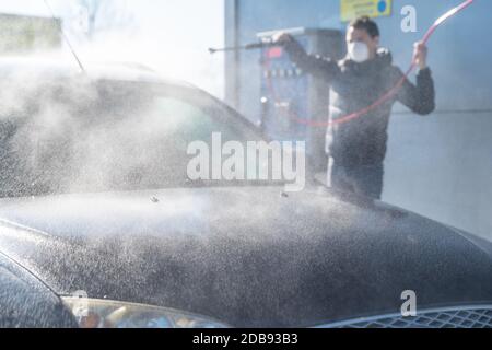 lavage de voiture par pression d'eau à la station libre-service. Un homme avec un respirateur sur le visage protège contre une épidémie de coronavirus. Banque D'Images