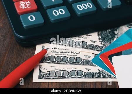 Une calculatrice noire, des factures de cent dollars, un stylo bille rouge vif et des cartes de crédit multicolores s'étalent sur une table en bois marron. Concept d'interdiction Banque D'Images