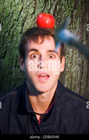 Vue frontale tête-et-épaules d'un jeune homme avec une pomme sur sa tête debout devant un arbre et regarde avec inquiétude la caméra avec sa mout Banque D'Images