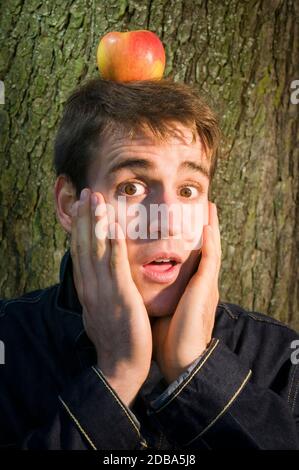 Vue frontale tête-et-épaules d'un jeune homme avec une pomme sur sa tête debout contre un arbre et regardant anxieusement à la caméra avec sa bouche op Banque D'Images
