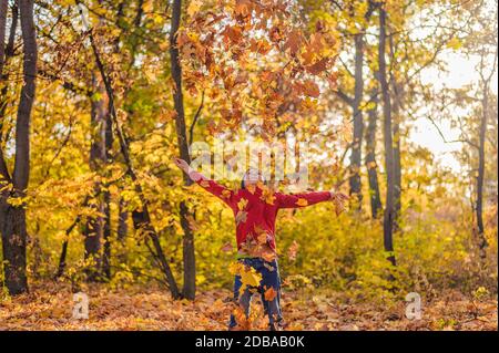 un heureux garçon dans un pull rouge jette un armful de feuilles jaunes dans la forêt d'automne Banque D'Images