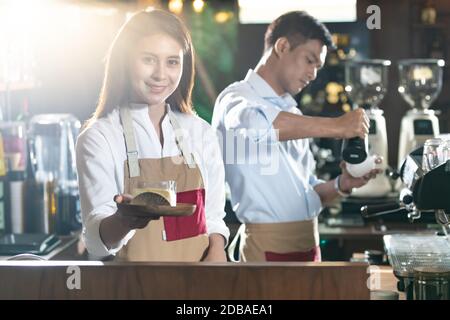 Portrait de la femme barista asiatique servant des gâteaux dans un café-restaurant au client avec d'autres collègues barista travaillant en arrière-plan. Utilisation pour petit jeu de barres Banque D'Images