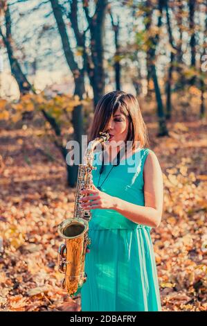 une jeune femme divine à poil sombre, vêtue d'une longue robe bleue, joue le saxophone alto sur le fond du feuillage tombé des arbres dans un autum jaune Banque D'Images