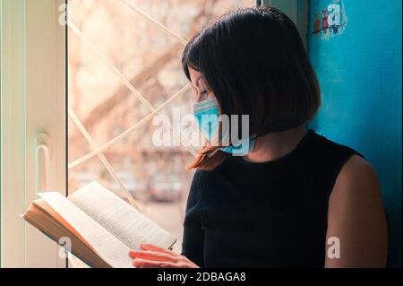 une fille à poil sombre dans une robe noire se trouve dans un masque chirurgical à la fenêtre de l'hôpital et lit un livre Banque D'Images