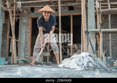 Maçon asiatique tenant une houe pour la pâte pour le sable et ciment pour bâtiments sur un fond de bâtiment de maison Banque D'Images