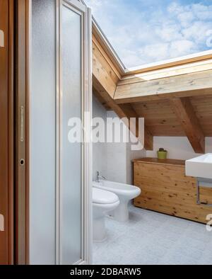 Salle de bains pratique et confortable avec toilettes, armoire et lavabo. Toit avec poutres apparentes et fenêtre du ciel. Banque D'Images