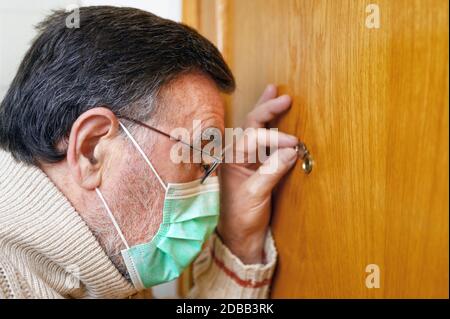 Concept de Covid-19. Restez à la maison. Auto-isolement pour prévenir la pandémie de coronavirus. Homme senior dans le masque vert de protection regarde à travers la péephole . Banque D'Images