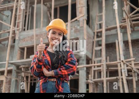 belle femme sous-traitant avec les pouces en haut portant un casque de sécurité dans le contexte de la construction de bâtiments Banque D'Images