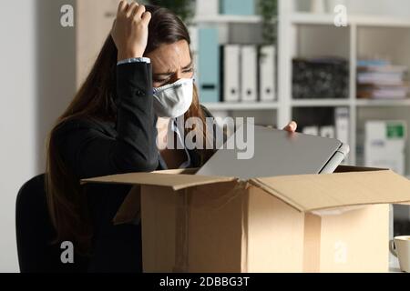 Femme exécutive au feu avec masque de protection pour l'emballage des effets personnels sur une boîte au bureau la nuit Banque D'Images