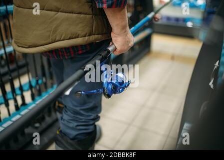 Le pêcheur à la ligne mâle tient la tige avec la bobine dans la boutique de pêche. Équipement et outils pour la pêche et la chasse au poisson, choix d'accessoires sur la vitrine en magasin, gains a Banque D'Images