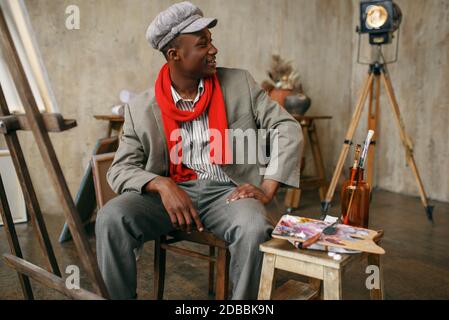 Un peintre masculin tendance en chapeau et foulard rouge pose au chevalet en studio d'art. L'artiste dessine sur son lieu de travail, des œuvres de maître créatives en atelier Banque D'Images