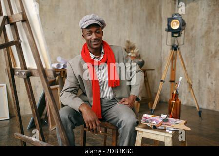 Un peintre masculin tendance en chapeau et foulard rouge pose au chevalet en studio d'art. L'artiste dessine sur son lieu de travail, des œuvres de maître créatives en atelier Banque D'Images