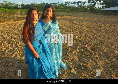 Deux adolescentes portant un sari bleu, doré, boucles d'oreilles, collier, nez avec des poils longs et foncés debout sur un champ agricole labouré Banque D'Images