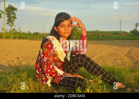 Gros plan sur une adolescente portant une robe traditionnelle colorée avec des poils longs et foncés assis sur l'herbe près d'une pelouse labourée champ et main sur la tête Banque D'Images