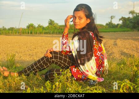 Gros plan sur une adolescente portant une robe traditionnelle colorée avec des poils longs et foncés assis sur l'herbe près d'une pelouse labourée champ et main sur la tête Banque D'Images