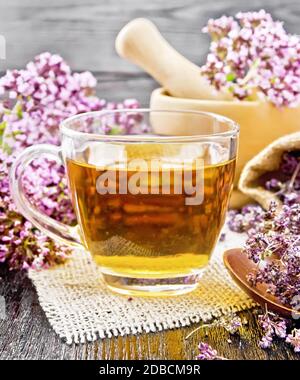 Thé à base de plantes origan dans une tasse de verre sur toile de jute, fleurs fraîches dans le mortier et sur la table, fleurs de marjolaine séchées dans un sac et une cuillère sur le dos de planche en bois Banque D'Images