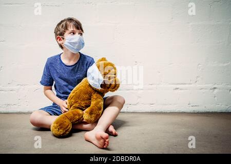 Un jeune garçon portant un masque tenant son ours en peluche pendant la pandémie de covid-19. Banque D'Images