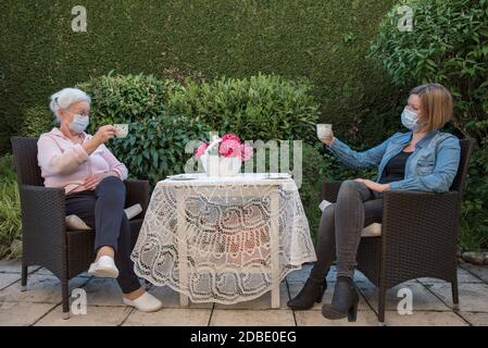 Femme et fille aînée avec masques de visage ayant un café dans le jardin à distance de sécurité Banque D'Images