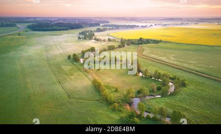 Printemps matin panorama rural aérien. Lever de soleil sur les champs de colza verts et jaunes fleuris. Nuages de brouillard et petite rivière avec arbres sur la rive. Bela Banque D'Images