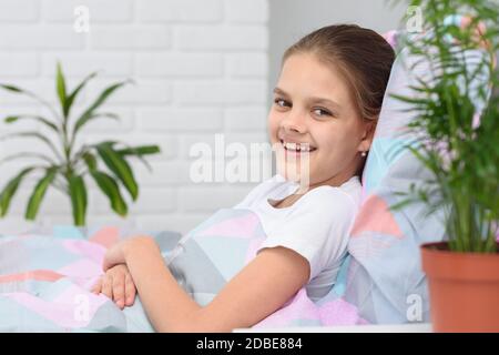 Une jeune fille heureuse de dix ans se trouve dans le lit et sourit dans le cadre. Banque D'Images