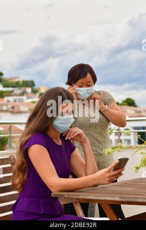 Femme asiatique mettant sur son masque chirurgical tandis qu'une femme hors foyer vérifie son téléphone sur un fond hors foyer. Concept de sécurité et d'amitié. Banque D'Images