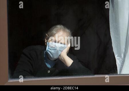 Une vieille grand-mère dans un masque de protection regarde la fenêtre à l'auto-isolation. Femme âgée en quarantaine à domicile. Banque D'Images