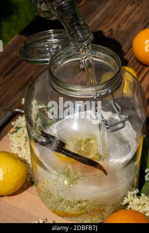 Versez la vodka dans le grand pot de conservation avec les fleurs de sureau fraîches, les oranges et les tranches de citron, le sucre et la gousse de vanille pour la liqueur de sureau Banque D'Images