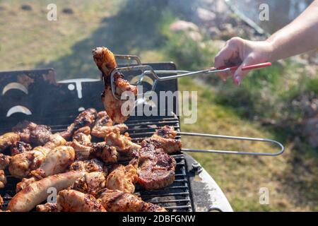 Une femme au gril à charbon de bois est de griller des steaks, des saucisses de gril et des ailes de poulet et à l'aide de pinces de barbecue. Mise au point sélective sur le gril contre un gr Banque D'Images